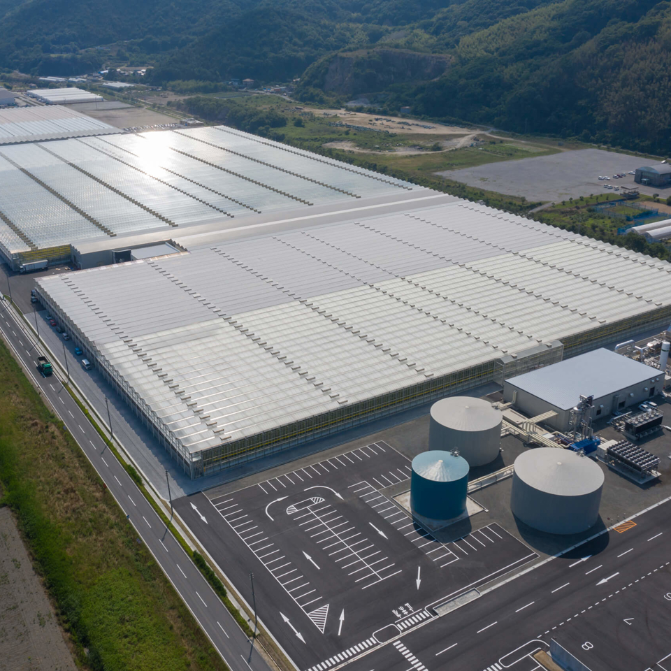 Aerial view of a large turnkey greenhouse complex in Kasaoka, Japan, featuring glass-covered structures, water tanks, and modern climate control facilities.