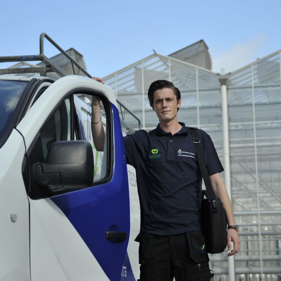 Technician standing next to a white and blue company van in front of a modern greenhouse, wearing a navy work uniform